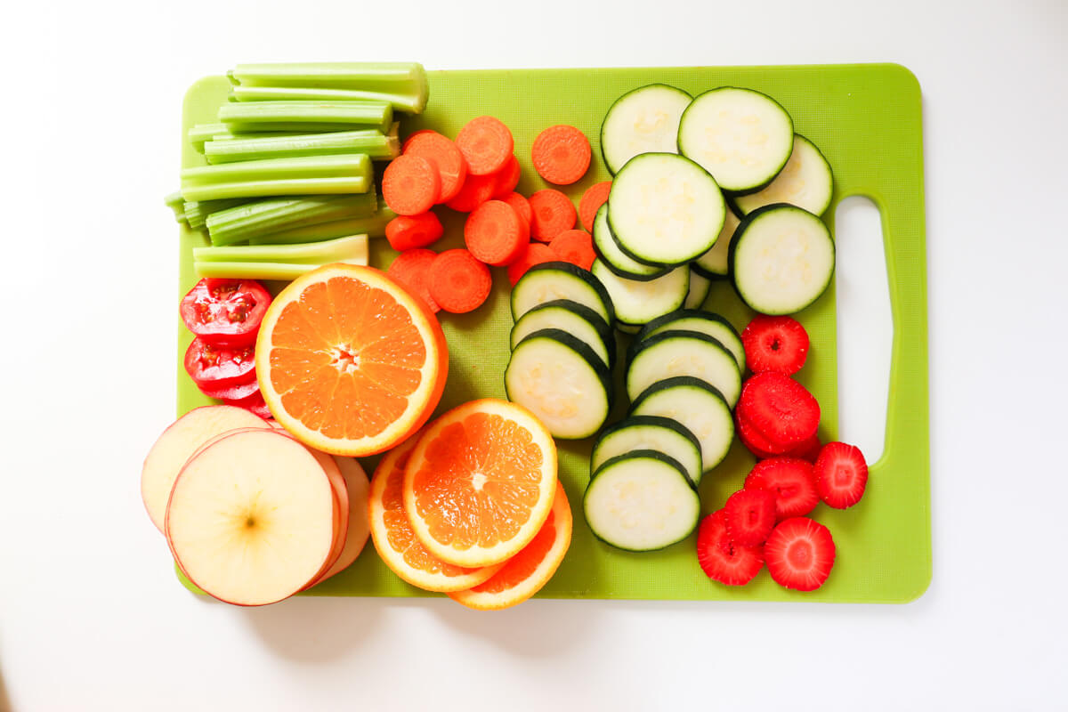 Cut up oranges, cucumbers, apples, celery sticks, carrots, strawberries on a green cutting board.
