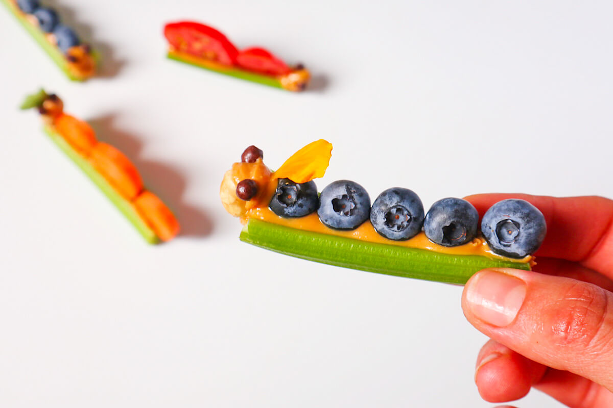 Ant on a log held by a hand made from celery sticks, peanut butter and blueberries.
