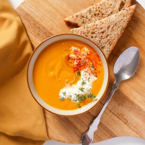 carrot and red lentil soup in a bowl topped with olive oil and feta and bread and a spoon next to it