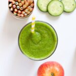 Top down view of a green smoothie in a glass with apple, cucumber and colored pencils on the side.