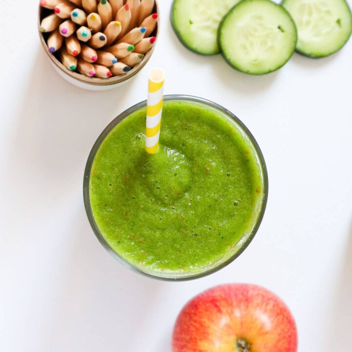 Top down view of a green smoothie in a glass with apple, cucumber and colored pencils on the side.